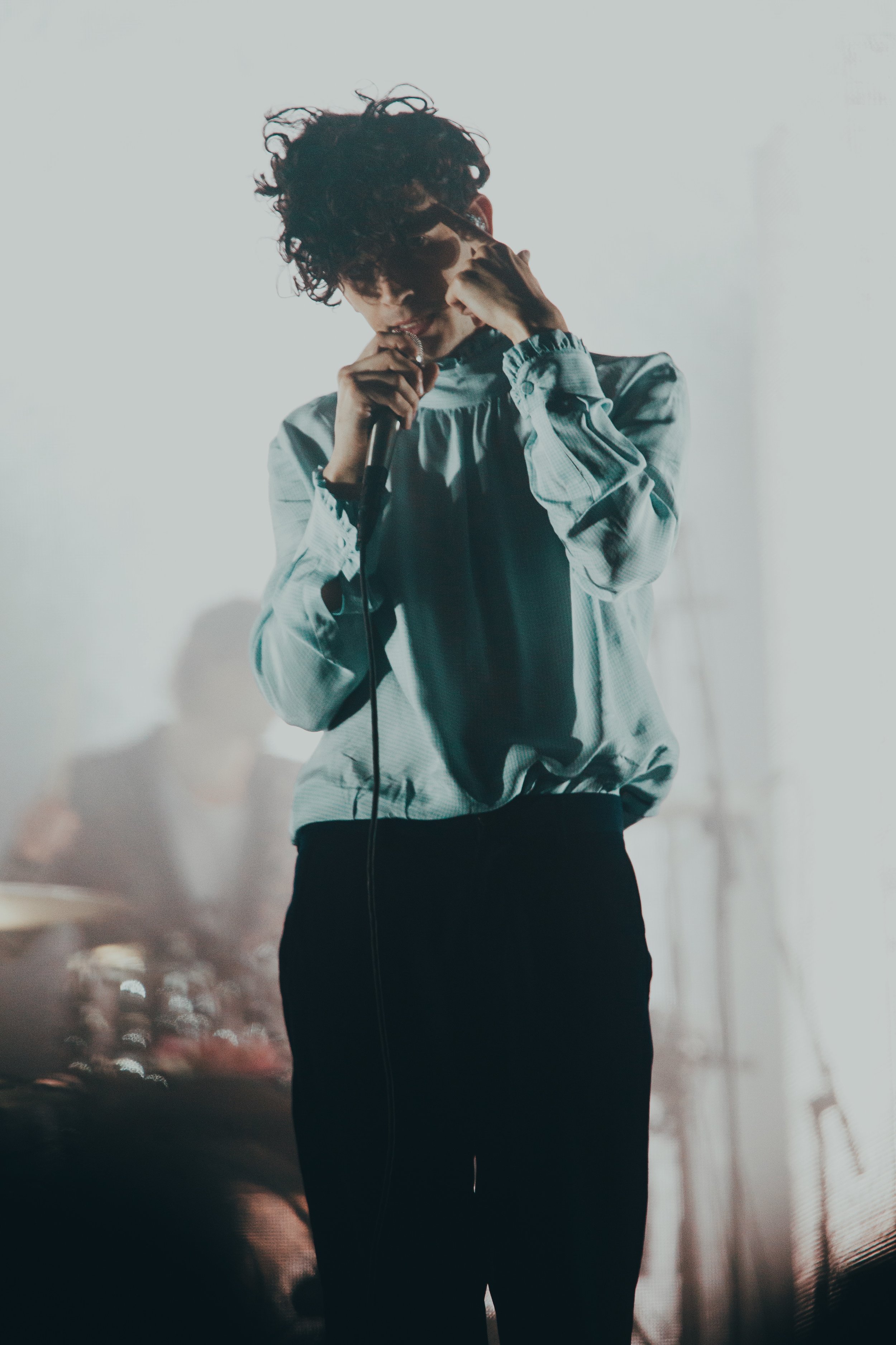 Matty, a white, male musician in his 20s, is wearing a ruffled teal blouse with sleek black trousers. His curly black hair casts his face in shadows and conceals his eyes. One hand clenches a microphone while he uses the index finger of the other to point to his forehead. This position correlates to a lyric in the song he was singing. Behind him, but obscured from view by the blurring of the background, is the band’s drummer.