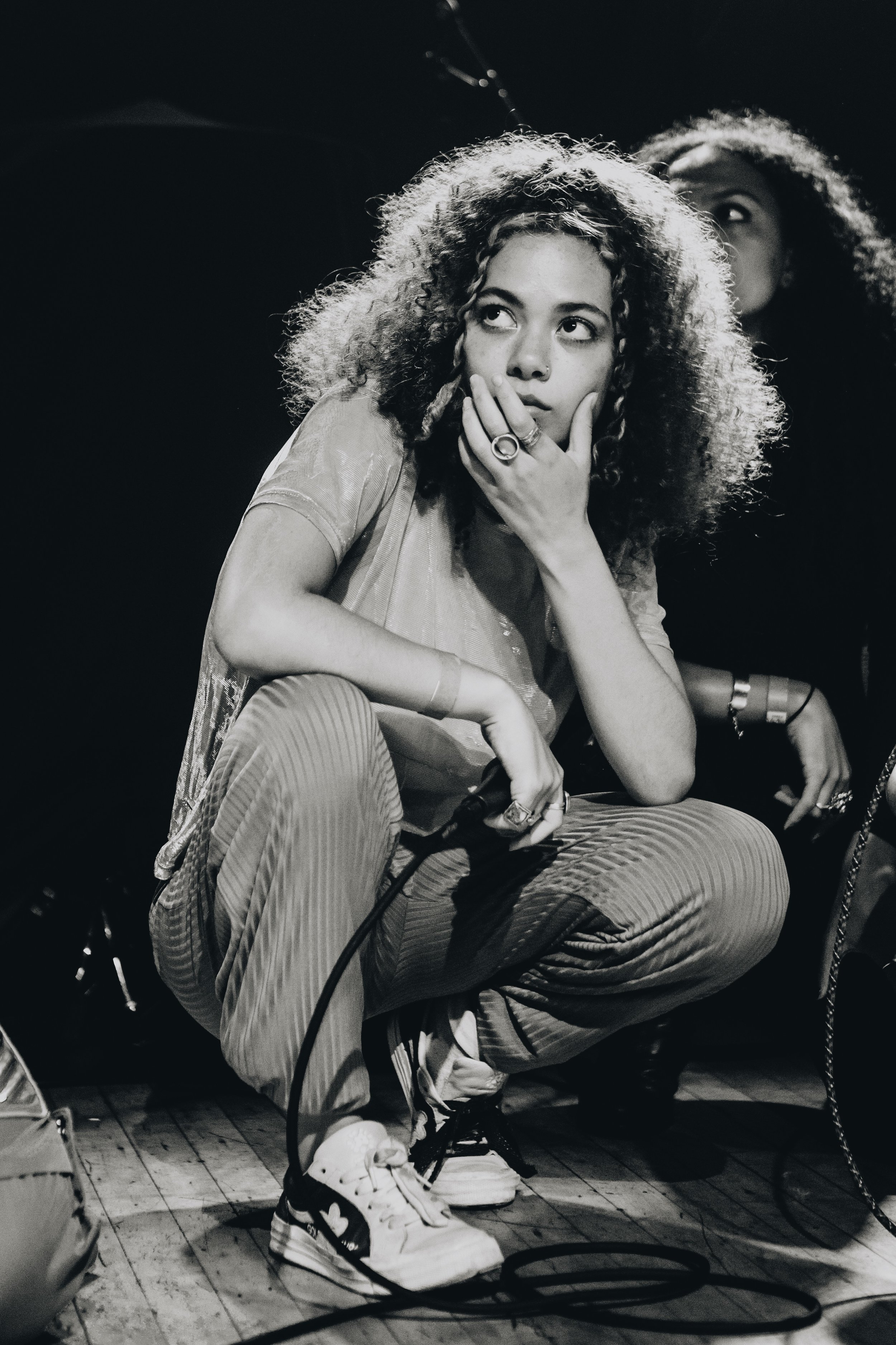 This black and white image depicts Isa, a member of the R&B collective MICHELLE. On the small stage of Warsaw she is joined by six other people. However, she is the sole subject of this image, crouched on stage and holding one hand up to her chin. This signifies a moment of contemplation, perhaps depicting Isa marveling at the audience (the largest MICHELLE has performed for to date).